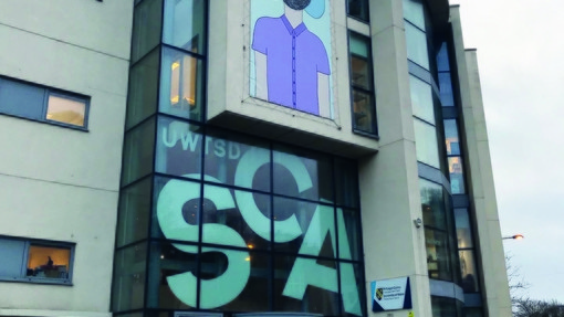 Image of Swansea College of Art Dynevor Building, a beige brick building with glass front with SCA in white writing with graphic image of boy in purple t-shirt